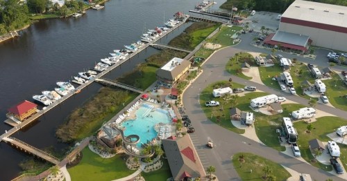 Aerial view of a marina with boats, a pool area, and RVs along a riverbank surrounded by greenery.