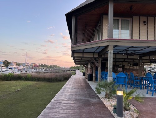 A waterfront restaurant with blue chairs, a wooden walkway, and boats in the background under a pastel sky.