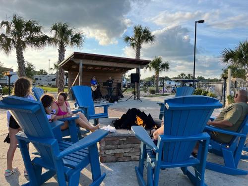 A group enjoys a live band by a fire pit, seated in blue chairs surrounded by palm trees and a scenic outdoor setting.
