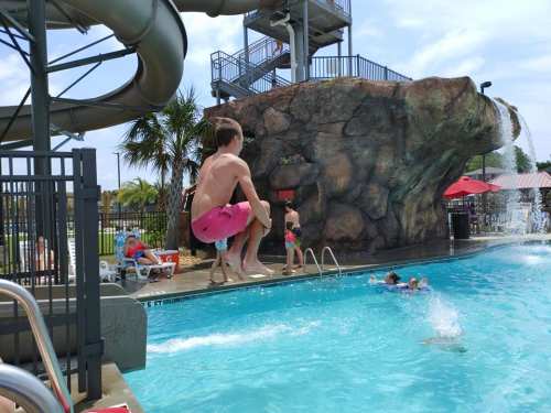 A boy in pink shorts jumps into a pool near a water slide, with children playing and a rock formation in the background.