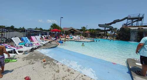 A sunny pool scene with people swimming, lounging on chairs, and a water slide in the background.