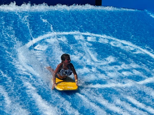A child rides a yellow boogie board on a wave simulator, splashing water against a blue background.