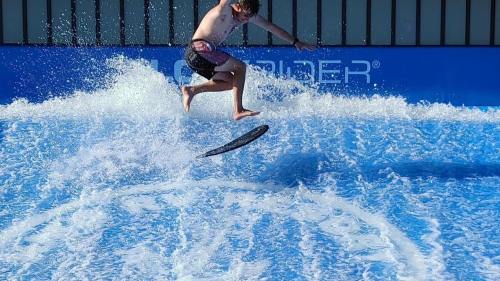 A young boy jumps off a surfboard, performing a trick on a wave simulator with splashing water around him.