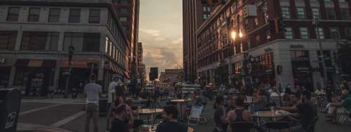 A bustling city street at sunset, with people sitting at outdoor tables and tall buildings lining the road.