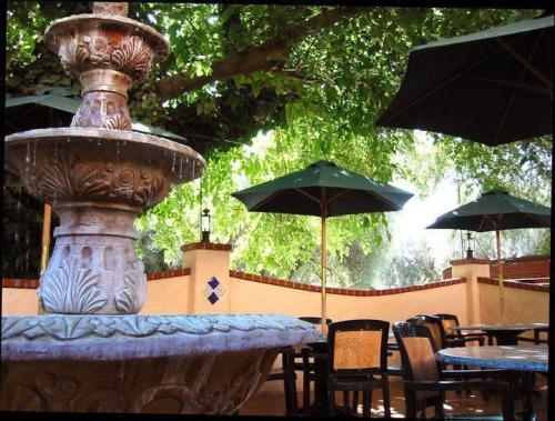 A decorative fountain surrounded by green trees and shaded tables with umbrellas in a cozy outdoor setting.