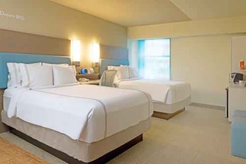 A modern hotel room featuring two beds, a desk, and a window with natural light. Decor is simple and inviting.
