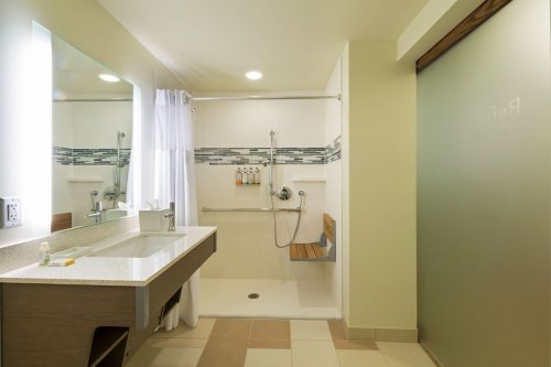 Modern bathroom with a walk-in shower, sink, and light-colored walls. Features a large mirror and accessible design elements.