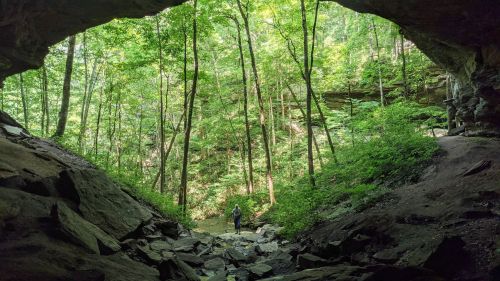 A person stands at the entrance of a cave, surrounded by lush green trees and rocky terrain.