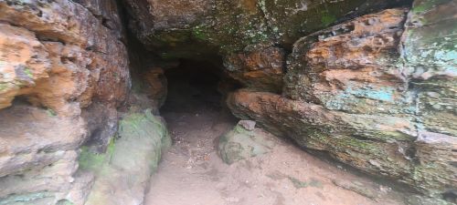 A rocky cave entrance with moss-covered walls and a sandy floor, leading into darkness.