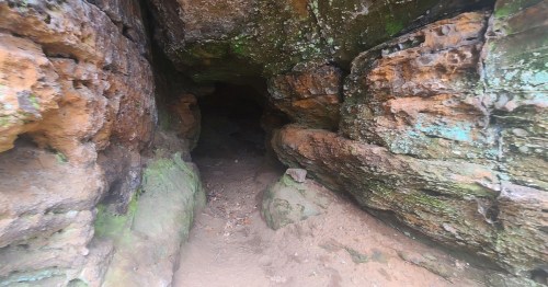 A rocky cave entrance with moss-covered walls and a sandy floor, leading into darkness.