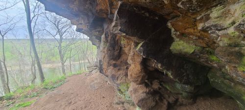 A rocky cave entrance with moss-covered stones, overlooking a green landscape and river in the background.