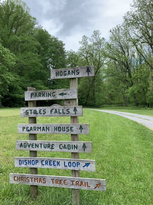 Wooden signpost with directional arrows for various trails and locations in a green, wooded area.