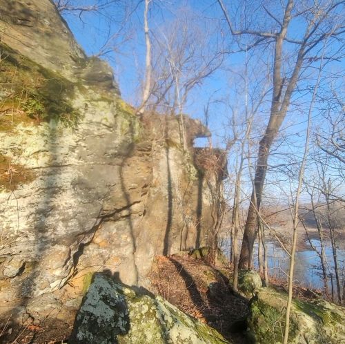 Rocky cliffside with trees and shadows, overlooking a river under a clear blue sky.