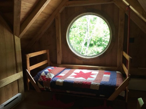 Cozy wooden bedroom with a round window overlooking greenery and a quilted bedspread featuring a star pattern.