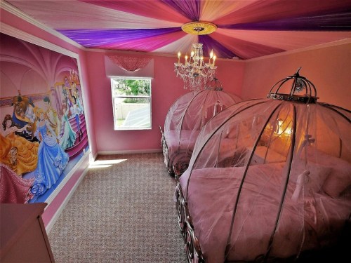 A whimsical pink bedroom with two canopy beds, a chandelier, and a princess-themed mural on the wall.