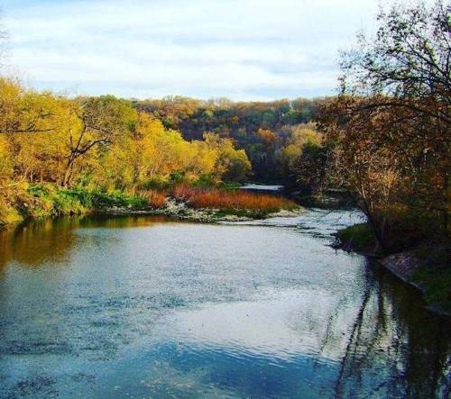 A serene river flows through a landscape of autumn trees, reflecting the colorful foliage in calm waters.