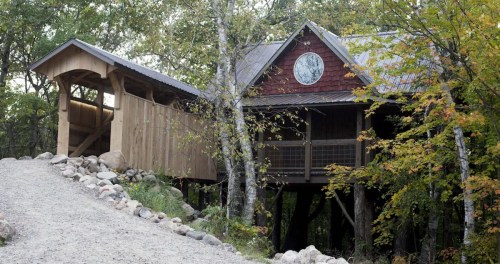 A wooden cabin on stilts surrounded by trees, with a stone pathway leading up to it and a clock on the front.