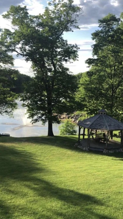 A serene lakeside view featuring a gazebo surrounded by lush green grass and trees under a partly cloudy sky.