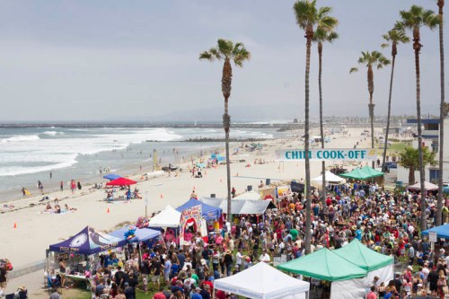 A crowded beach scene with palm trees, tents for a chili cook-off, and waves in the background under a cloudy sky.