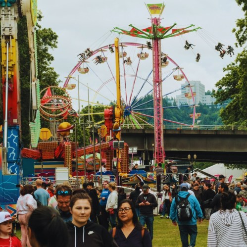 A lively fair scene with colorful rides and a crowd of people enjoying the festivities under a cloudy sky.