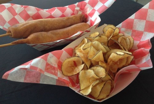 Two corn dogs on skewers and a serving of crispy potato chips in red and white checkered baskets.
