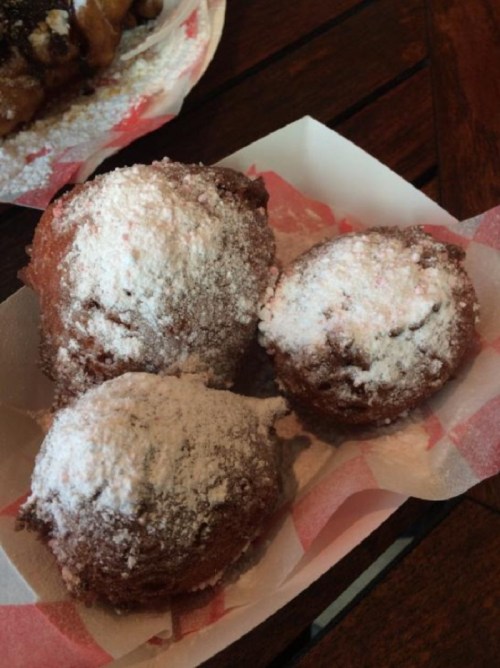Three powdered sugar-dusted fried pastries on a paper tray, with a rustic wooden background.