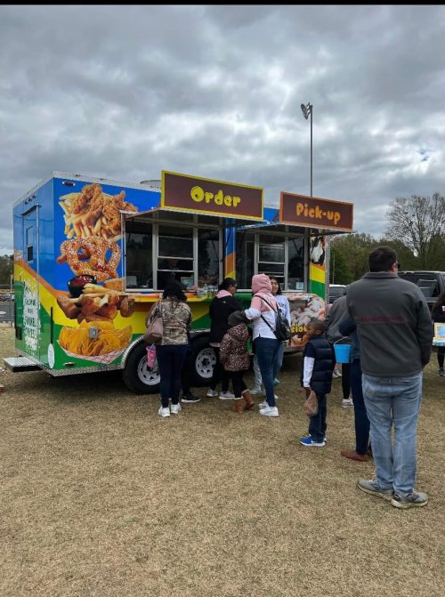A colorful food truck with "Order" and "Pick-Up" signs, surrounded by a line of people on a grassy field.