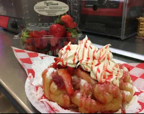 A delicious funnel cake topped with whipped cream, strawberries, and strawberry drizzle, served on a checkered paper.