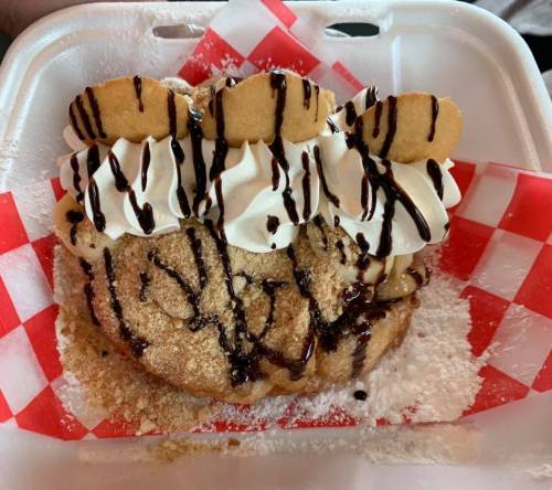 A dessert featuring a large, fried dough base topped with whipped cream, chocolate drizzle, and cookie pieces.