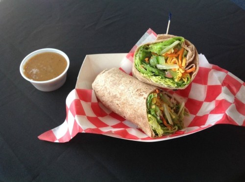 Two wrapped veggie sandwiches on a checkered paper tray, accompanied by a small cup of sauce.