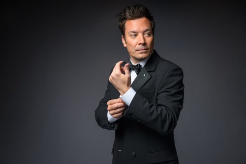 A man in a tuxedo adjusts his cufflink, looking confidently to the side against a dark background.