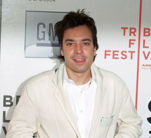 A man with tousled hair smiles at an event, wearing a light jacket over a white shirt against a festival backdrop.