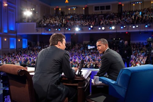 A talk show host interviews a guest on stage in front of a large audience, with bright lights and cameras present.