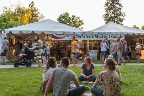 A lively outdoor art exhibition with people browsing tents and a group sitting on the grass enjoying the event.