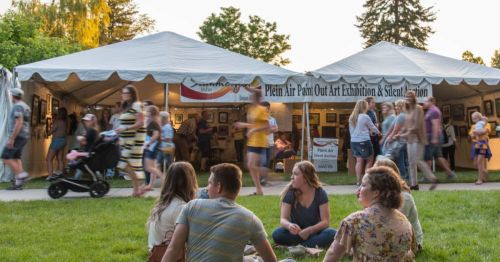 A busy outdoor art exhibition with tents, people browsing artwork, and a group sitting on the grass in the foreground.