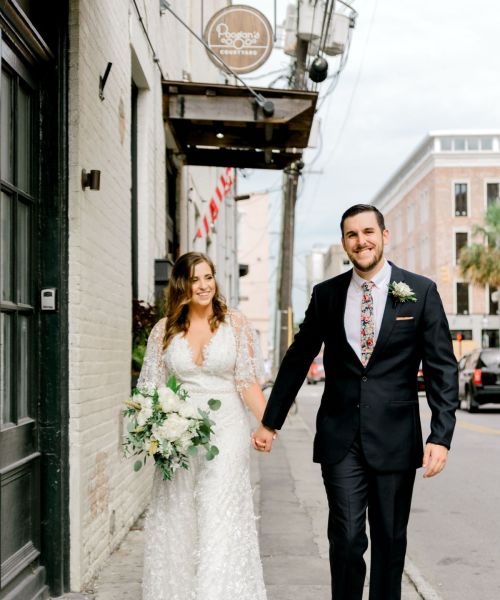 A bride and groom walk hand in hand down a charming street, smiling, with the bride holding a bouquet of flowers.