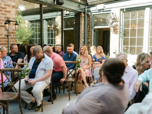 A group of people seated indoors, chatting and enjoying a gathering in a stylish venue with large windows.