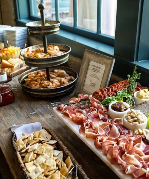 A buffet table featuring shrimp, cured meats, cheeses, crackers, and condiments, with a window in the background.