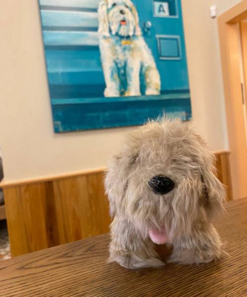A fluffy dog figurine sits on a table, with a painting of a similar dog on the wall behind it.