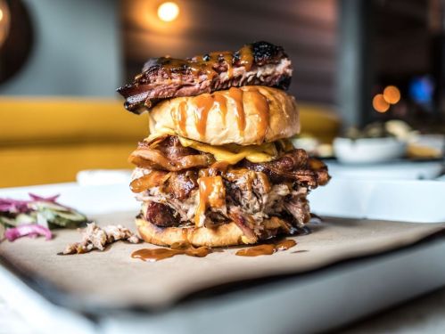 A towering burger stacked with pulled pork, cheese, and barbecue sauce, served on a plate with onions on the side.