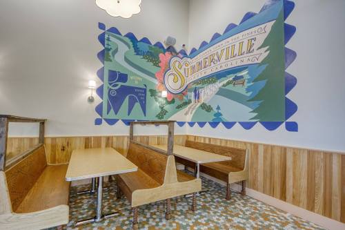 A cozy restaurant corner featuring a colorful mural of Summerville, South Carolina, with wooden booths and patterned flooring.