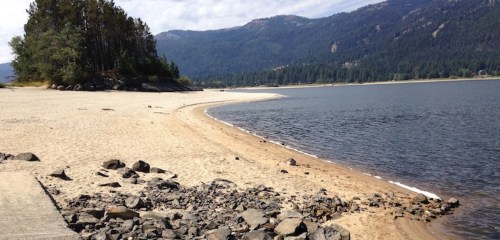 A sandy beach curves along a calm lake, surrounded by trees and mountains under a partly cloudy sky.