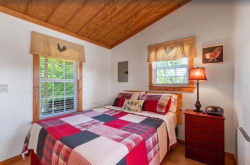 Cozy bedroom with a wooden ceiling, a quilted bedspread, and a bedside lamp next to a window with decorative curtains.