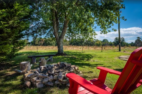 A red Adirondack chair beside a stone fire pit under a large tree, with a grassy field in the background.