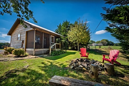A cozy cabin with a grassy yard, two red chairs by a fire pit, and trees under a clear blue sky.