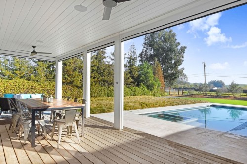 A modern outdoor patio with a dining table, pool, and lush greenery in the background under a clear blue sky.