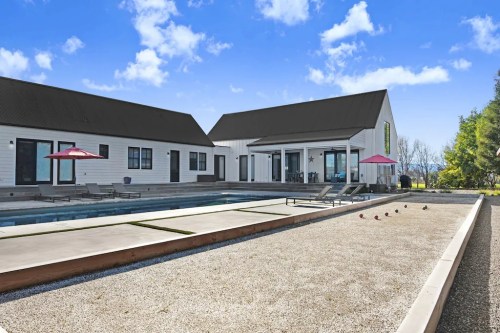 Modern home with a pool and bocce court, surrounded by greenery and blue skies. Pink umbrellas add a pop of color.