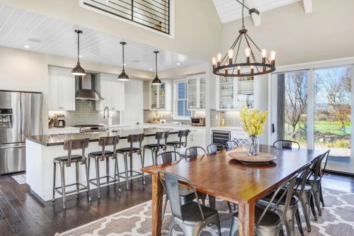 Bright, modern kitchen and dining area with a large wooden table, bar stools, and large windows overlooking greenery.