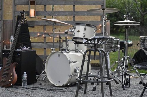 A drum set and bass guitar are set up on a gravel surface, with a stool and wooden backdrop in a garden setting.