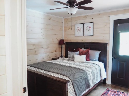 Cozy bedroom with a wooden bed, decorative pillows, and a ceiling fan, featuring light wood paneling and a door.